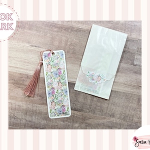 May include: A white bookmark with a floral design and a pink tassel. The bookmark is packaged in a clear cellophane bag with a floral sticker.