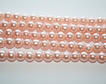 May include: Rows of light peach-colored, round beads. The beads are strung together, creating multiple strands. The beads have a glossy finish, reflecting light. The background is a plain white.