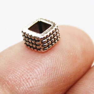 May include: Silver square spacer bead with a textured surface. The bead has a hole in the center for stringing.