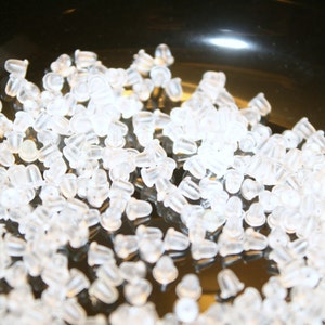 May include: A close-up shot of a collection of clear, silicone earring backs. The earring backs are small, with a rounded shape and a hole for the earring post. They are scattered across a dark, reflective surface.