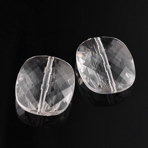 May include: Two clear, faceted beads with a square shape and a hole running through the center. The beads are transparent, allowing light to pass through, and are set against a black background. These beads are suitable for jewelry making.
