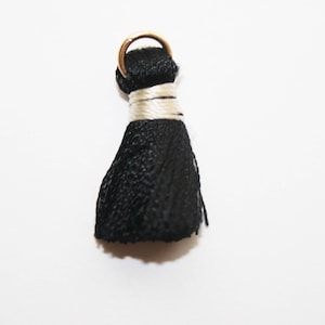 May include: A black tassel with a gold-colored ring at the top, wrapped with a cream-colored thread. The tassel is made of a woven material and hangs down from the ring.