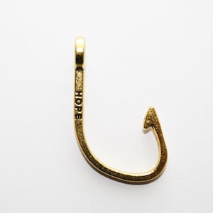 May include: A gold-colored fish hook pendant with the word "HOPE" engraved on the shaft. The hook is curved with a pointed tip and a loop at the top for a chain or cord. The pendant is set against a white background.