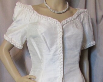May include: A white two-piece outfit with a braided trim. The top has a V-neck and short sleeves. The skirt is a-line and falls to the knees. The outfit is made of a silky fabric.