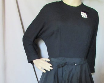 Könnte beinhalten: Ein schwarzes Vintage-Kleid mit langen Ärmeln und einem weiten Rock. Das Kleid hat ein figurbetontes Oberteil und einen ausgestellten Faltenrock. Eine dekorative Brosche ist auf der oberen Brust befestigt. Das Kleid ist ein klassischer Stil.