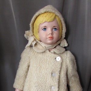 May include: A vintage doll wearing a white faux fur coat with a hood and a white scarf. The doll has blonde hair and blue eyes.