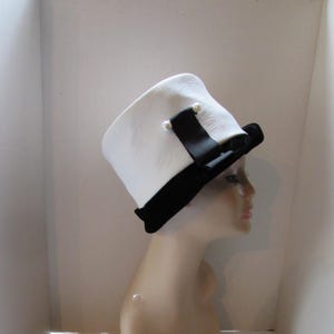 May include: A white hat with a black brim and a black ribbon, adorned with two small pearl-like embellishments. The hat is displayed on a mannequin head, showcasing its design and shape. A classic accessory.