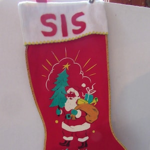 May include: Red Christmas stocking with a gold trim and a white cuff. The cuff has the word "SIS" embroidered in red. The stocking features a vintage illustration of Santa Claus carrying a bag of gifts and a Christmas tree. The stocking also has the words "Merry Christmas" embroidered in green.