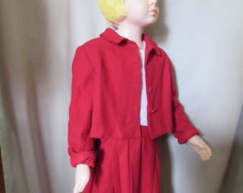 May include: A red skirt suit with a matching jacket and skirt. The jacket has a Peter Pan collar and long sleeves. The skirt is knee-length and has pleats.