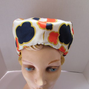 May include: A white satin hat with a geometric pattern of orange, red, and navy blue. The hat is shaped like a dome and has a smooth, shiny finish.