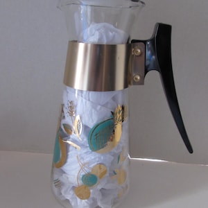May include: A vintage glass pitcher with a gold band and black handle. The clear glass is decorated with gold and teal fruit and floral designs. The pitcher is filled with white tissue paper.