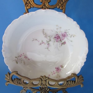 May include: A white porcelain bowl with a delicate floral pattern in shades of pink and purple. The bowl is displayed on a gold metal stand.