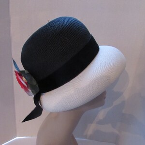 May include: A two-tone hat with a black crown and a white brim. A black velvet band encircles the crown, and a decorative floral accent with red, white, and blue elements is attached. A black ribbon hangs from the side.