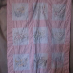 May include: A pink and white patchwork quilt with nine embroidered squares. Each square features a different hand-stitched design, including baskets and figures. The quilt's border is a solid pink, and the squares are white.