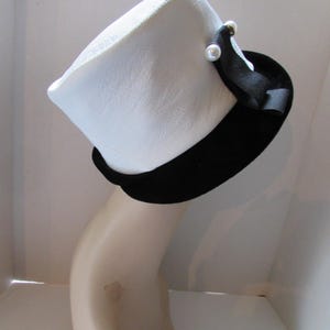 May include: A white hat with a black band and a black ribbon accent. The hat is adorned with two small, faux pearl embellishments. The hat is displayed on a mannequin head.