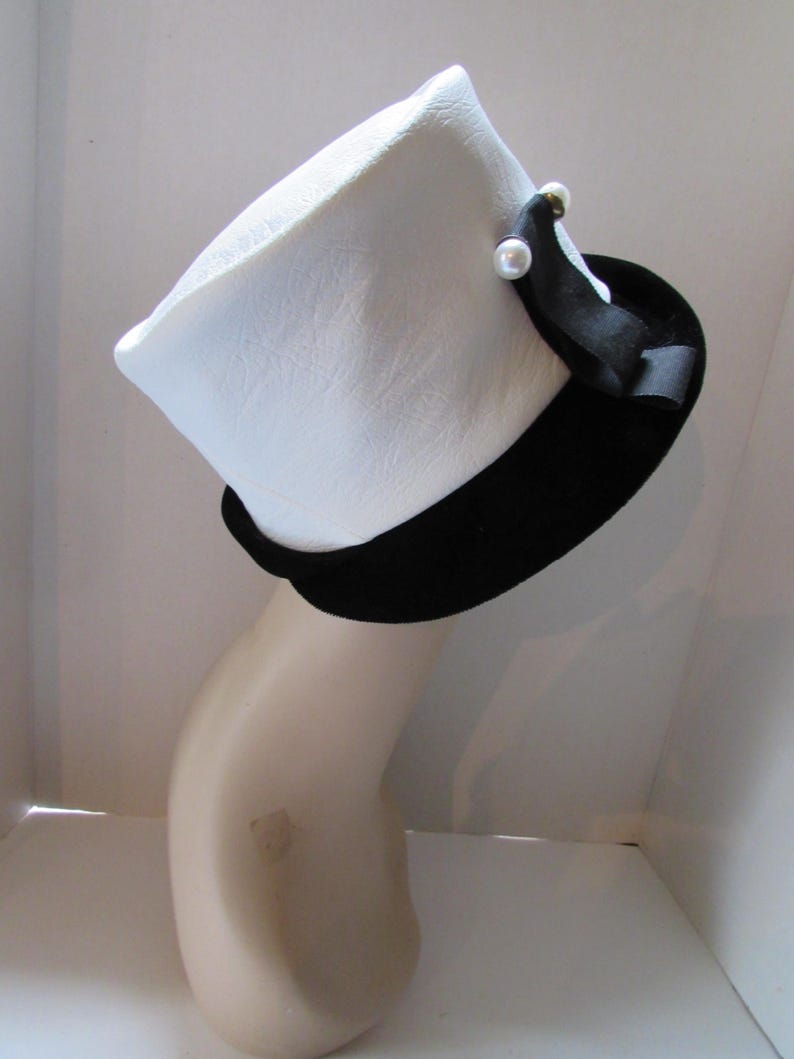 May include: A white hat with a black band and a black ribbon accent. The hat is adorned with two small, faux pearl embellishments. The hat is displayed on a mannequin head.