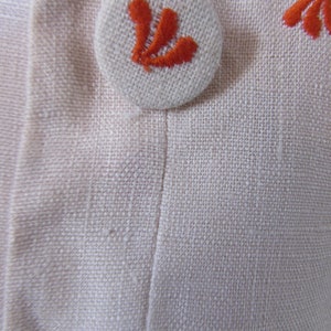May include: A white button with an orange embroidered design. The button is sewn onto a white fabric background.