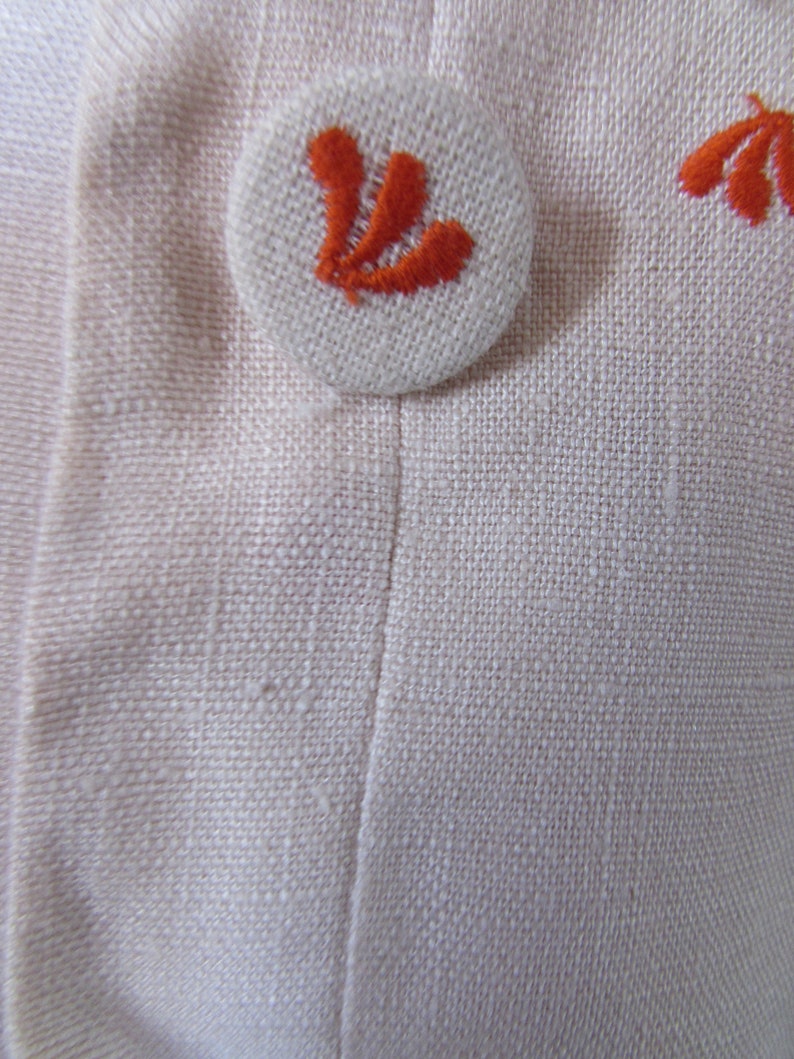May include: A white button with an orange embroidered design. The button is sewn onto a white fabric background.