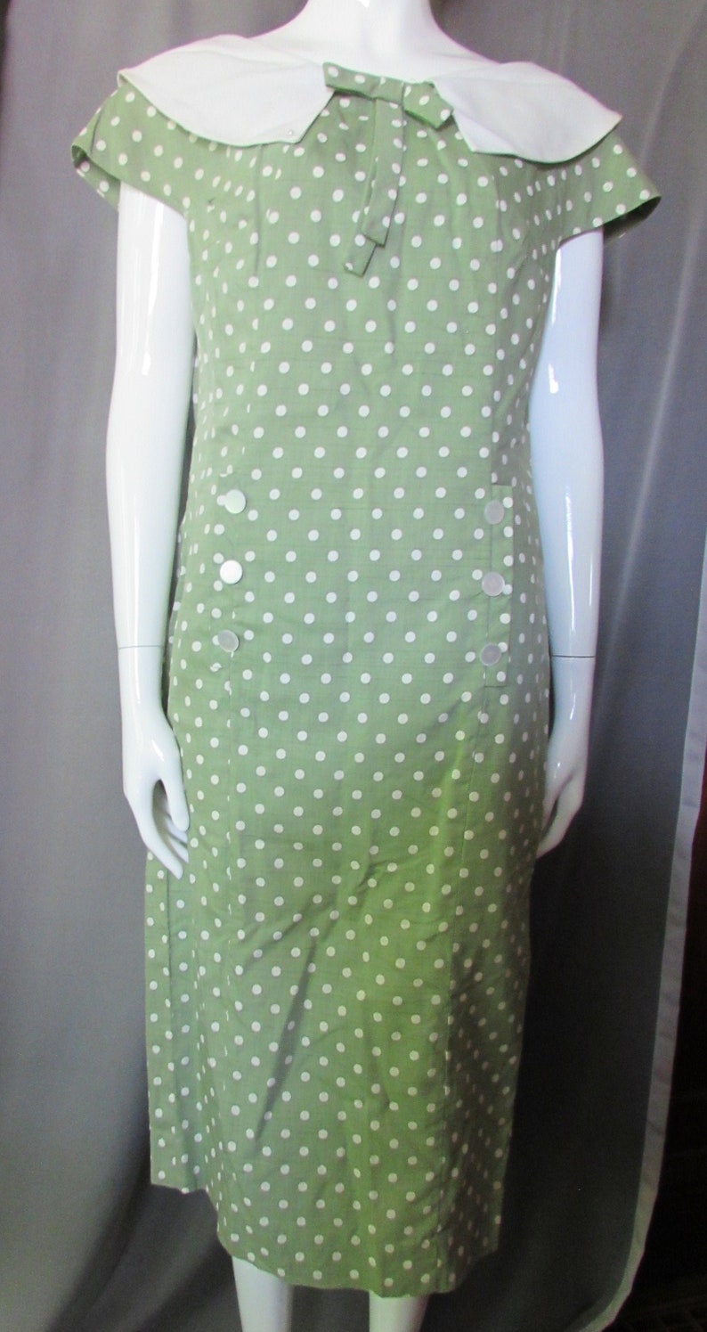May include: A green dress with white polka dots and a white collar. The dress has a bow at the neckline and buttons down the front.