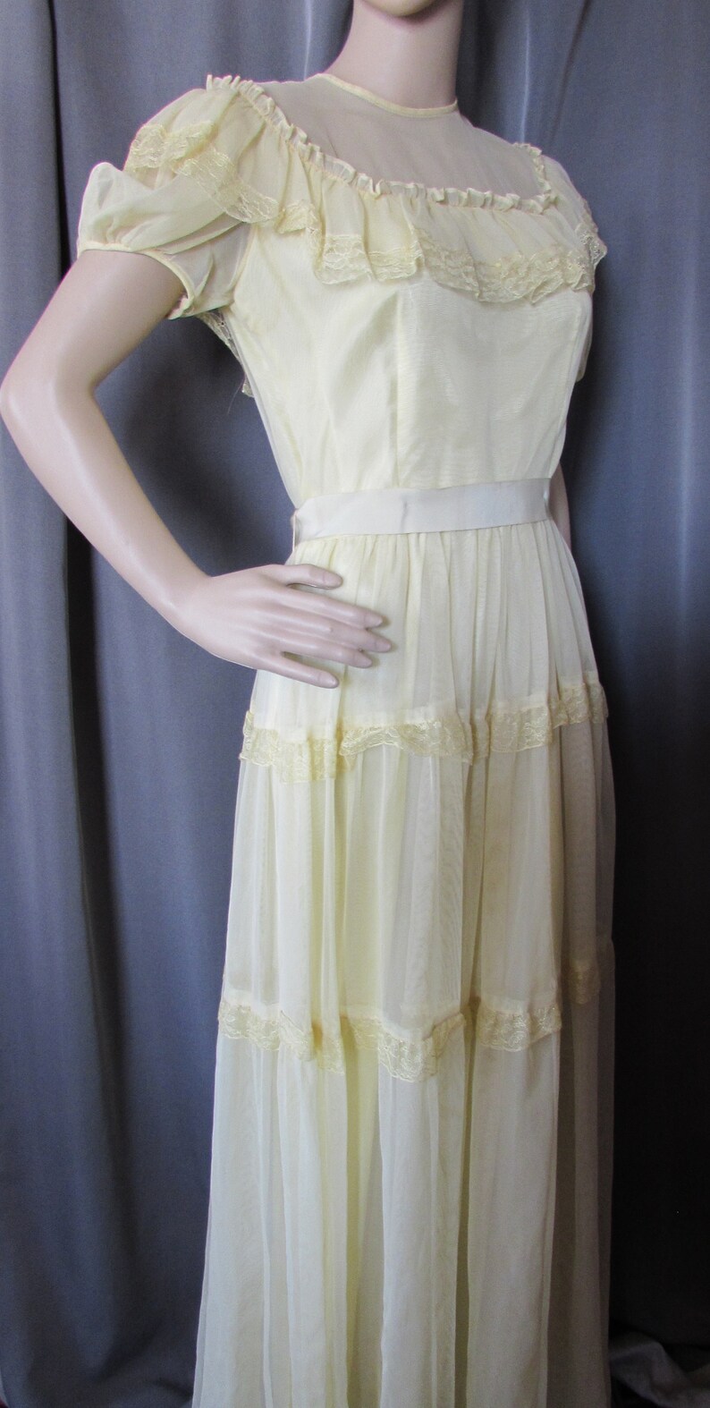 May include: A pale yellow, floor-length dress with a lace trim and a satin sash. The dress has a ruffled neckline and short sleeves.