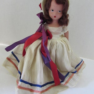 May include: A vintage doll with brown hair and a white dress with blue and red trim. The doll is wearing a white bonnet with a red flower.