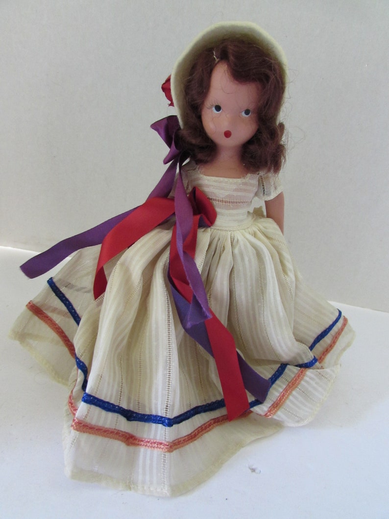 May include: A vintage doll with brown hair and a white dress with blue and red trim. The doll is wearing a white bonnet with a red flower.