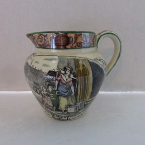 May include: Cream-colored ceramic pitcher with a green rim and handle. The pitcher features a detailed illustration of a historical scene with figures in period clothing. The design includes a dark blue and black background with text at the base.