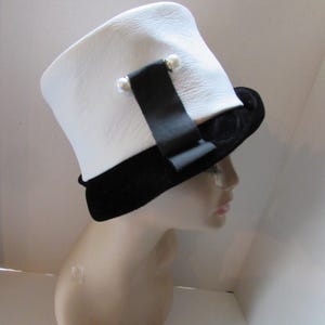 May include: A white hat with a black velvet brim and a black ribbon detail. The hat features two small pearl accents on the ribbon. The hat is displayed on a mannequin head.