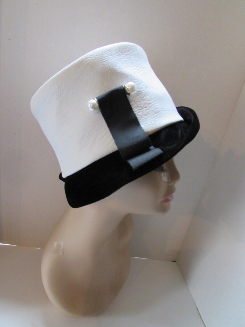 May include: A white hat with a black velvet brim and a black ribbon detail. The hat features two small pearl accents on the ribbon. The hat is displayed on a mannequin head.