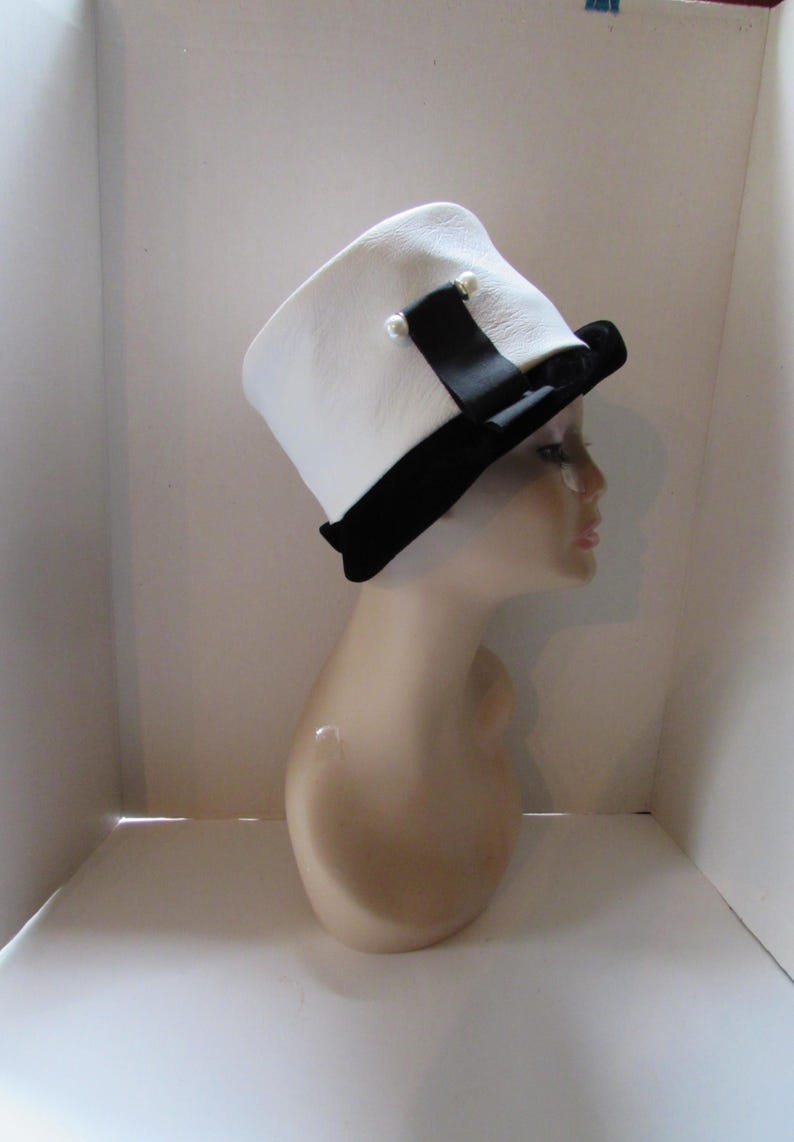 May include: A white hat with a black brim and a black ribbon, adorned with two small pearl-like embellishments. The hat is displayed on a mannequin head, showcasing its design and shape. A classic accessory.
