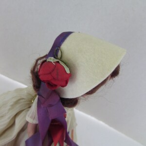 May include: A vintage doll with brown hair wearing a white felt hat with a purple trim and a red rose. The doll is wearing a white dress with purple and red ribbons.
