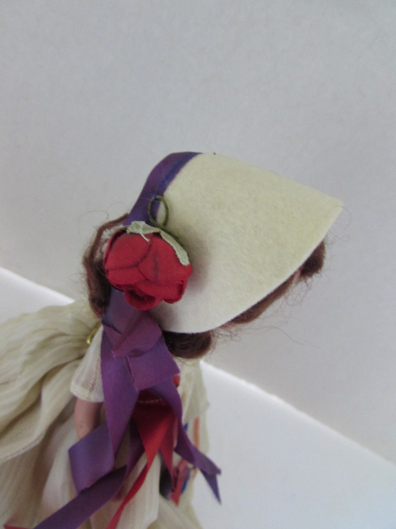 May include: A vintage doll with brown hair wearing a white felt hat with a purple trim and a red rose. The doll is wearing a white dress with purple and red ribbons.