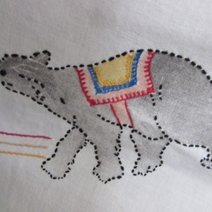 May include: Embroidered gray bear with a red, blue, and yellow saddle on a white fabric background.