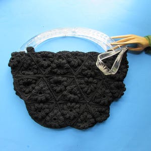 May include: Black crocheted handbag with a clear, curved handle. The bag features a floral pattern and a clear, decorative accent. The handle is attached to a wooden hand with a green ribbon. The bag is set against a blue background.