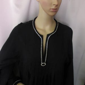 May include: A black long-sleeved tunic featuring a split neckline adorned with a row of clear rhinestones that form a circular accent at the base. The cuffs are also embellished with a matching single row of clear rhinestones along the edges.