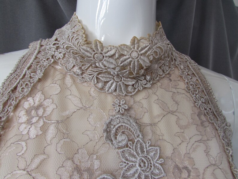 May include: A close-up of a beige lace halter top with a high neckline. The lace is delicate and features a floral pattern.