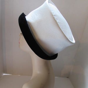May include: A white hat with a black brim is displayed on a mannequin head. The hat has a unique, angular shape and a textured surface. The black brim adds a contrasting accent to the hat's design. This hat is a fashion accessory.