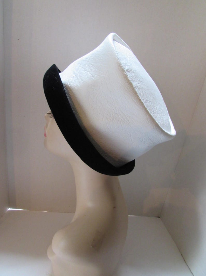 May include: A white hat with a black brim is displayed on a mannequin head. The hat has a unique, angular shape and a textured surface. The black brim adds a contrasting accent to the hat's design. This hat is a fashion accessory.