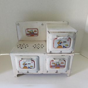 May include: A vintage white toy stove with the words "Pretty Maid" on the front. The stove has two burners, two drawers, and a small oven. Each section has a colorful illustration of a woman cooking.
