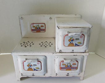 May include: A vintage white toy stove with the words "Pretty Maid" on the front. The stove has two burners, two drawers, and a small oven. Each section has a colorful illustration of a woman cooking.