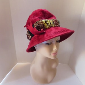 May include: A red felt hat with a wide brim and a feather trim. The hat is decorated with a band of feathers in shades of brown, yellow, and green.