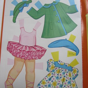 May include: A vintage paper doll page featuring cut-out clothing and accessories. Includes a green coat with blue trim, a pink tutu, a floral dress, and hats. The page is open, showing the colorful illustrations.