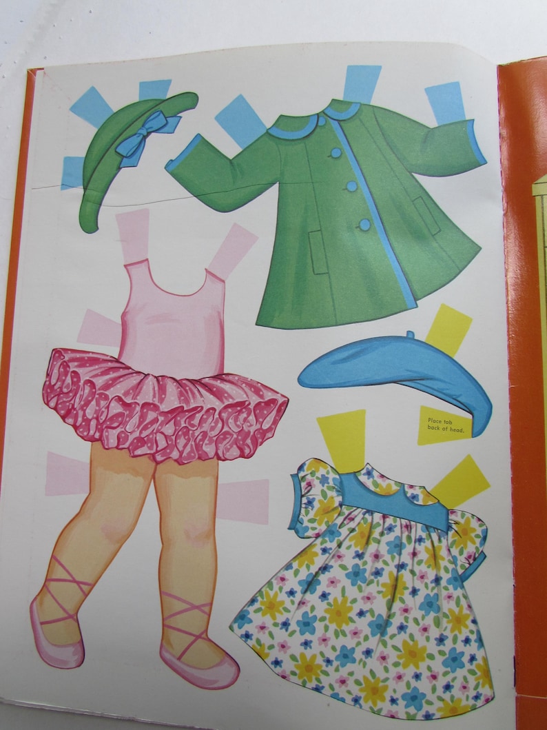 May include: A vintage paper doll page featuring cut-out clothing and accessories. Includes a green coat with blue trim, a pink tutu, a floral dress, and hats. The page is open, showing the colorful illustrations.