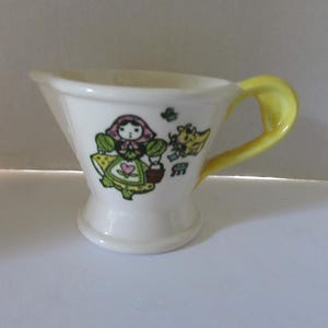 May include: A small, white ceramic cup with a yellow handle. The cup features a cartoon illustration of a girl with a basket and a dog. The handle and the illustration are in yellow, green, pink, and black.