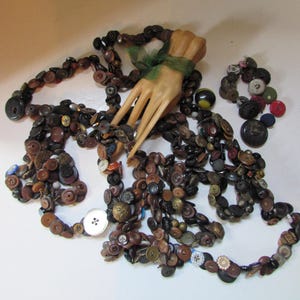 May include: A long string of assorted buttons in various shades of brown, black, and metallic tones, featuring a variety of sizes and textures. A wooden hand model is positioned in the center, tied with a thin green ribbon, surrounded by buttons.