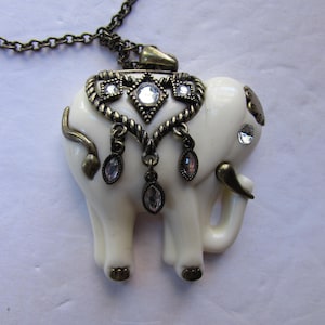 May include: A white elephant pendant necklace with a gold chain and rhinestone accents. The elephant is detailed with a gold trim and has a jeweled design on its back.