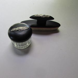 May include: An antique vanity set featuring a black powder jar with a silver filigree accent and a matching ink blotter. The ink blotter has a black handle and a beige absorbent pad.