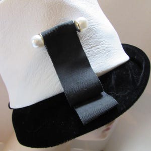 May include: A white hat with a textured surface, a black velvet brim, and a black ribbon detail. The ribbon is secured with two pearl-like embellishments. The hat is a classic style, suitable for various occasions.