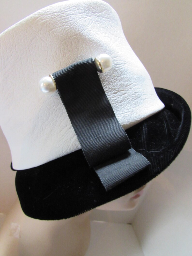 May include: A white hat with a textured surface, a black velvet brim, and a black ribbon detail. The ribbon is secured with two pearl-like embellishments. The hat is a classic style, suitable for various occasions.