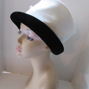 May include: A white hat with a black brim. The hat has a slightly curved top and a black band around the base. A small, white pearl-like embellishment is visible on the side. The hat is displayed on a mannequin head.